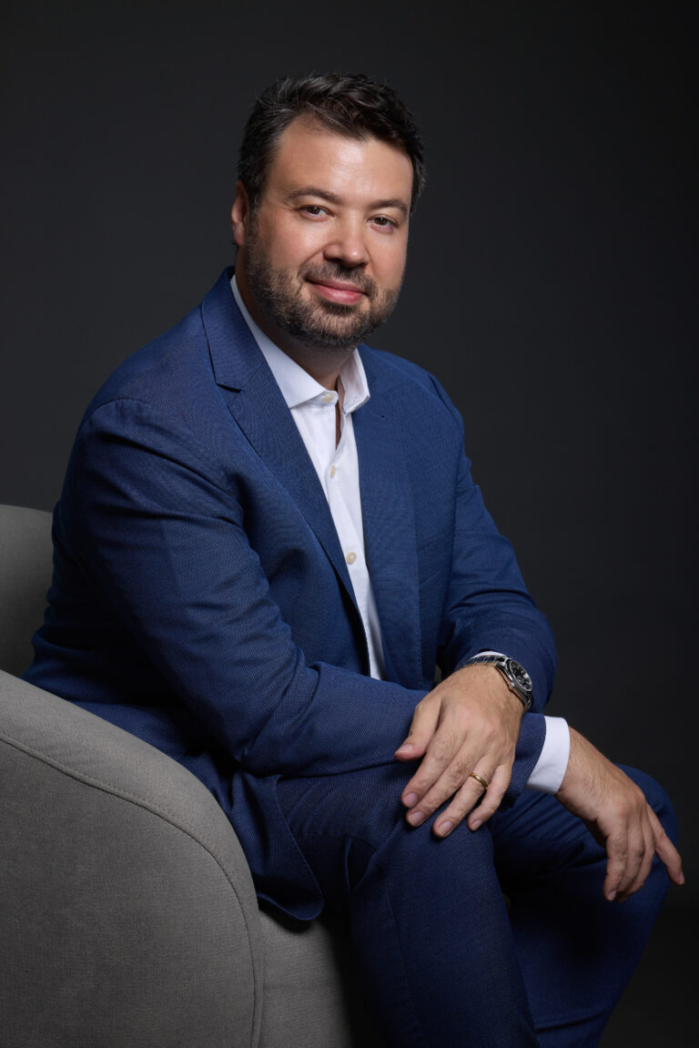 Retrato de estúdio de Marcelo Vitorino sentado em poltrona, de terno azul, sorrindo
