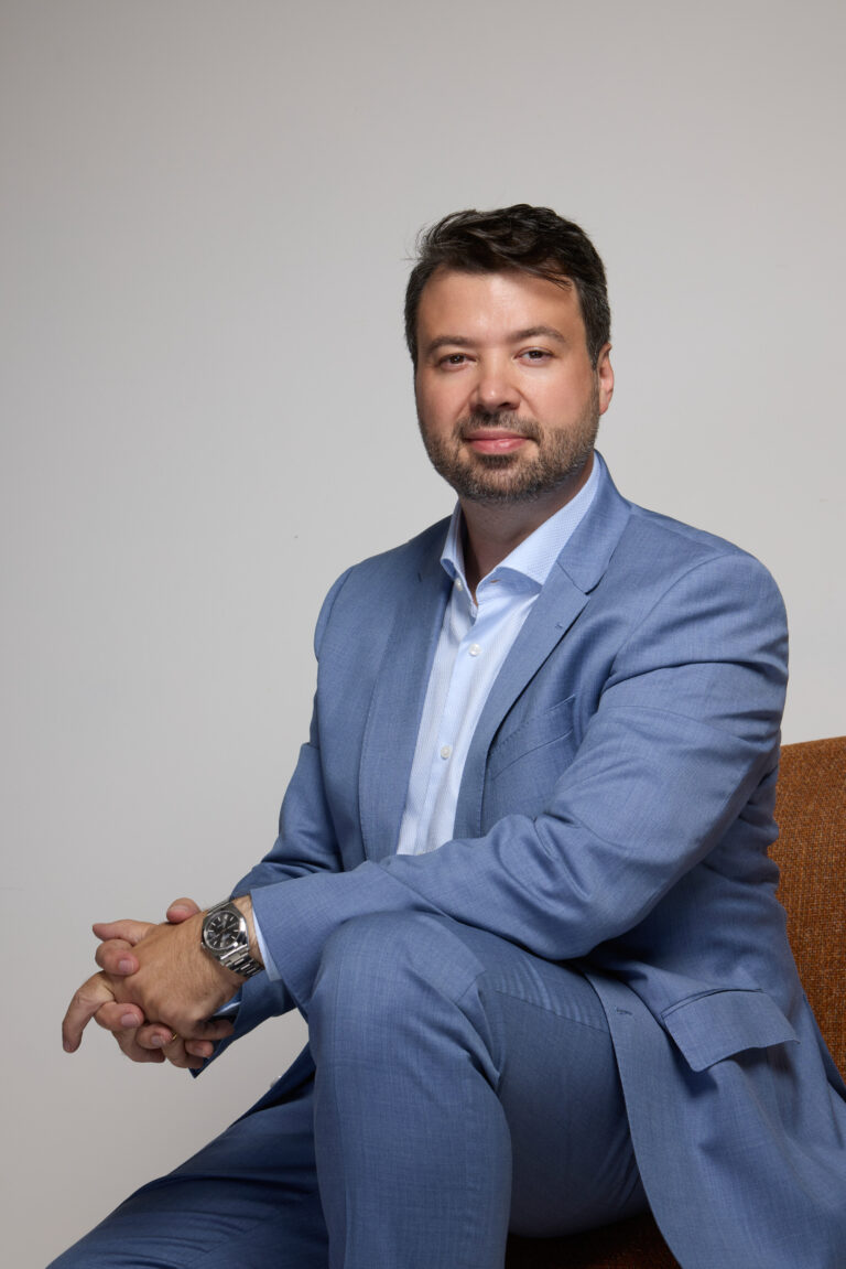 Retrato de estúdio de Marcelo Vitorino sentado, de terno azul-claro, sobre fundo neutro