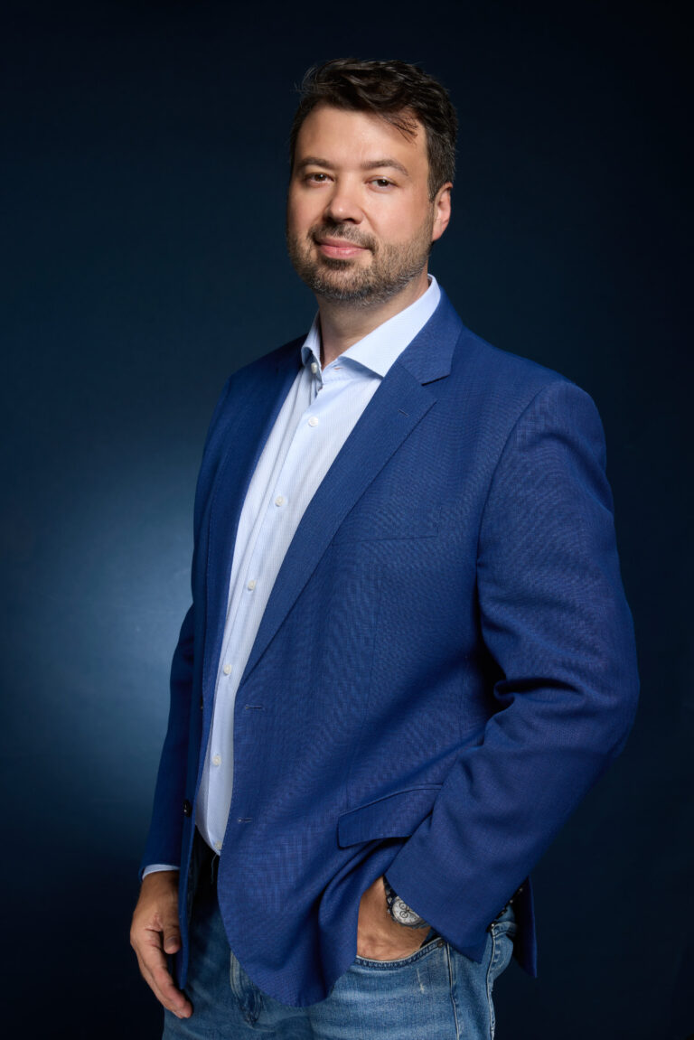 Retrato de estúdio de Marcelo Vitorino em pé, de blazer azul e camisa azul-clara, sobre fundo azul escuro